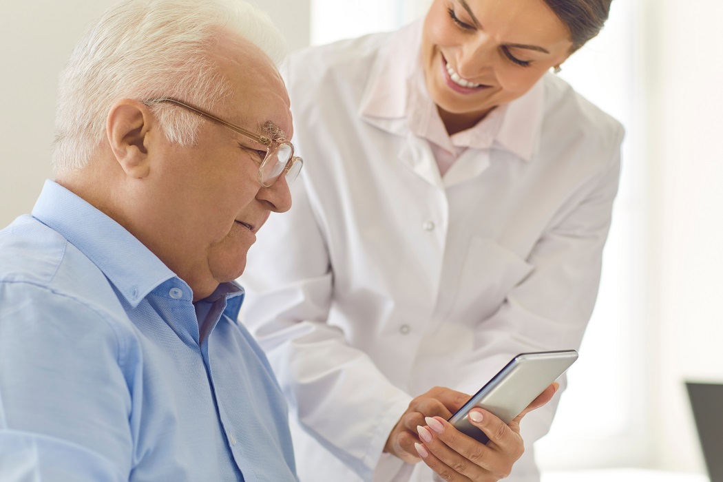 Smiling nurse teaching senior man to use healthcare app or showing photo of his grandchildren