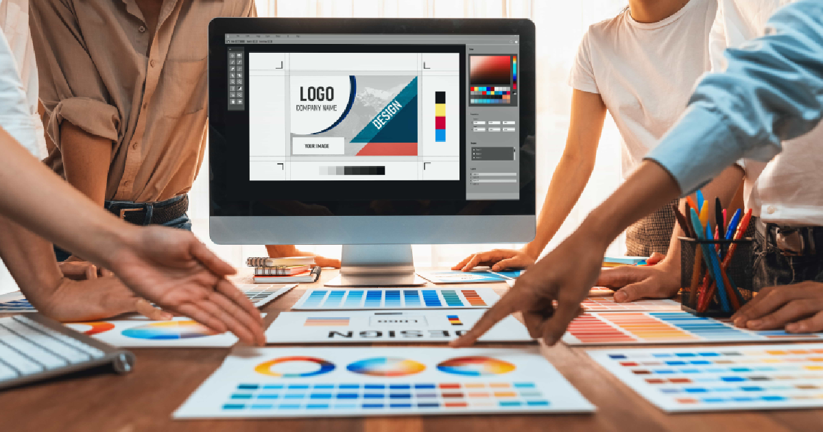 A group of people collaborating around a desk covered with color palettes, design prints, and markers, with a computer screen displaying a logo design in graphic design software.